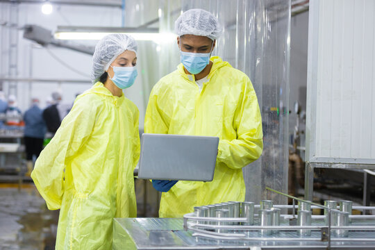 Two factory workers in protective uniforms and face masks inspect canned food production line with laptop in manufacturing. Food safety, quality control, hygiene, industrial inspection concept.