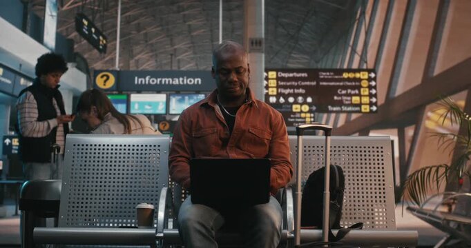 African American Man Sits in Terminal Hall, Concentrated on Laptop Screen, Waiting For Departure, With Bags and Coffee Nearby. Digital Nomad Lifestyle, Telecommuting, Global Connectivity of Traveler.