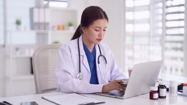 Asian adult woman doctor medical professional wearing lab coat stethoscope working laptop computer desk healthcare office clinic research patient records online consultation
