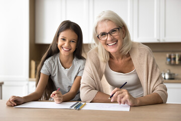 Fototapeta premium Portrait of senior granny and little cute granddaughter drawing pictures with colored pencils, spend time together on favourite hobby. Older relative help with task, teach schoolgirl to paint at home