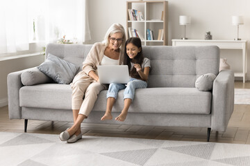 Obraz premium Middle-aged woman with little granddaughter do online shopping at home, sit on sofa in cozy living room with laptop, choose goods, buying e-commerce services via marketplace platform or website apps
