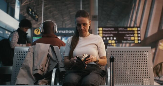 Woman Sits in Departure Hall, Holding Physical Boarding Pass While Checking Details on Smartphone, Having Already Prepared Luggage and Taken Off Coat. Concept of Airline Apps, and Passenger Security.