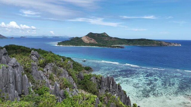 Push-in flyby drone shot over Ile Island highlighting dramatic limestone cliffs and vibrant shallow and deep blue waters with Patoyo Island in the distance, Philippines