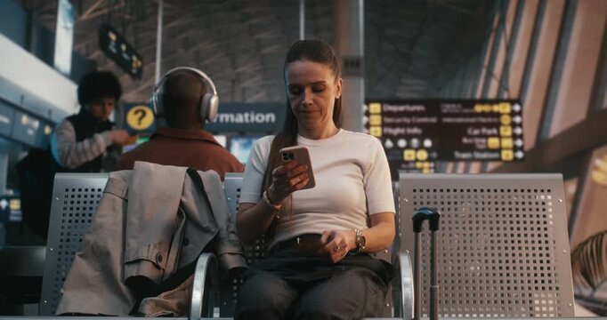 Female in T Shirt Finishes Preparations By Scanning Ticket With Smartphone. Woman Glances at Flight Information Display, Picks Up Trench Coat and Suitcase, and Confidently Walks Toward Boarding Area.