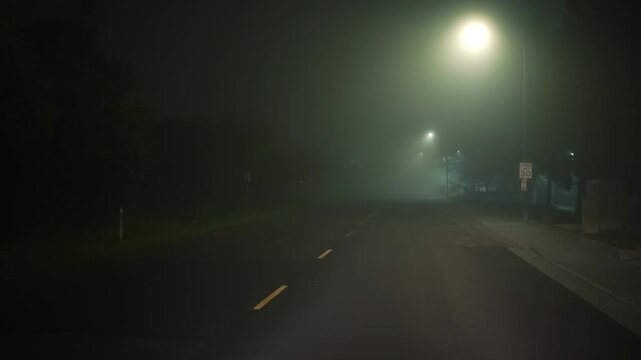 Driving along empty foggy night street with streetlights, forward motion along the road through dense fog creates atmosphere of night travel low visibility and quiet urban driving