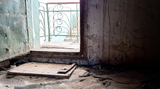 Dusty doorway with wooden panel and cracked walls leading to balcony with decorative metal railing, capturing aged textures, daylight, and atmosphere of abandonment