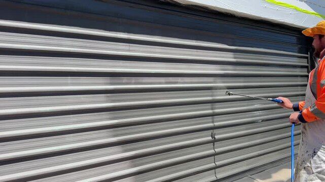 Worker Using Paint Sprayer to Repaint Metal Garage Door Exterior