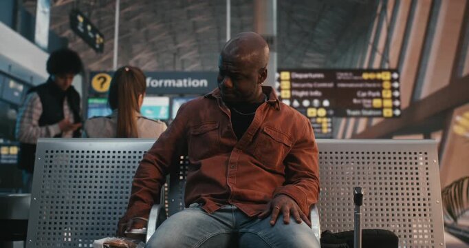 Man Takes Break From Journey Have Quick Snack. Sitting in Airport Waiting Area With Luggage Nearby, Male Unwraps Sandwich and Drinks Coffee in Paper Cup. Authentic Atmosphere of Long Transit Hours.