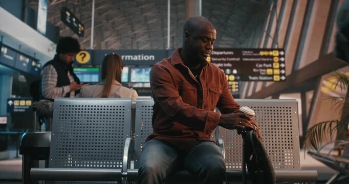 Young Black Man Arrives at Airport Waiting Area, Sets Down Luggage, Immediately Checks Smartphone. Modern Habit of Staying Connected, Checking Flight Updates, Chatting With Friends Before Departure.