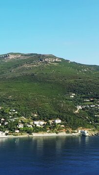 Scenic view of a lush green hillside with scattered houses overlooking the calm blue sea in Erbalunga, Corsica, under a clear sky.