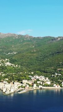 Aerial view of Erbalunga village nestled between lush green hills and the calm blue sea under a clear sky. Picturesque Mediterranean scenery with traditional buildings along the shoreline.