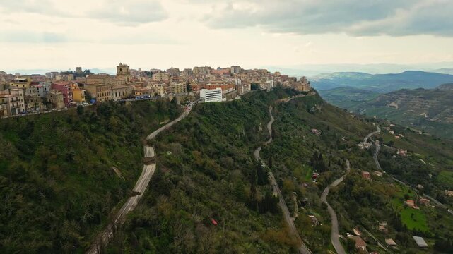 Aerial view of Enna, Sicily showcasing the scenic hillside cityscape with winding roads, lush greenery, and historical architecture under a cloudy sky
