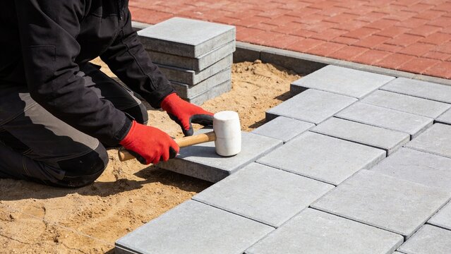 Construction worker laying down interlocking concrete pavers on a sandy surface with a rubber mallet for a new patio or walkway