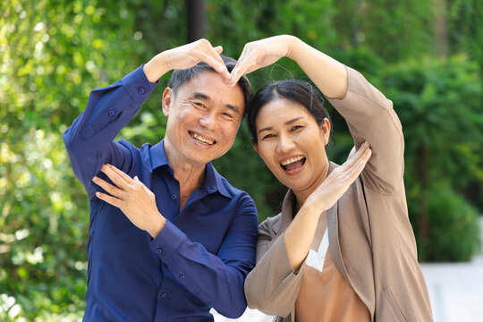 Happy and lovely married Asian senior couple, old casual husband and wife in urban park with good quality of life, making Asian style big heart body gesture.