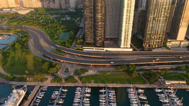Stunning aerial footage of Chicago downtown featuring luxury yacht marina and multilevel highway interchange with skyscrapers bathed in warm golden hour sunlight near waterfront park.