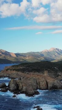 Dramatic rocky coastline with a historic Genoese tower overlooking the blue Mediterranean Sea near Cargese, Corsica, with mountains in the background under a partly cloudy sky.