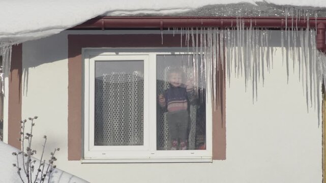 a child looks out the window of a house in winter, Long icicles on the roof of the house and a child in the window