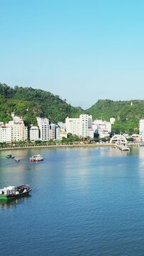 Scenic view of boats floating near the waterfront with green hills and modern buildings in Cat Ba Island, Vietnam on a sunny day.