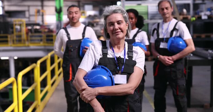 Confident senior female supervisor holding a blue hard hat and looking at the camera with her multi ethnic industrial team standing behind her in a modern manufacturing facility