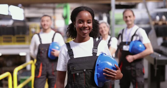 Confident young african american woman in overalls holding a blue hard hat and smiling at the camera, with her diverse team of multicultural colleagues standing proudly in the background