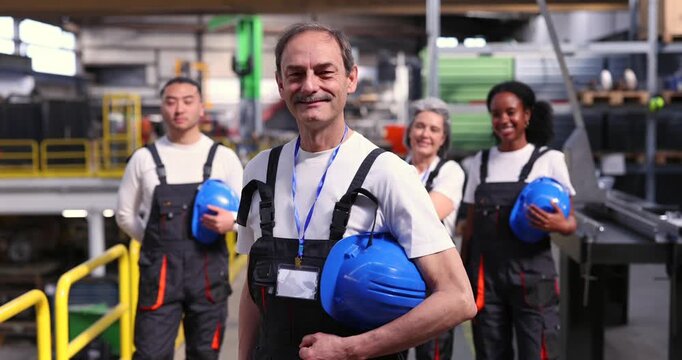 Senior male leader smiling at the camera holding a blue hard hat with his multi ethnic team of factory workers standing confidently in the background inside an industrial warehouse facility