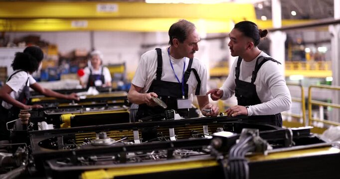 Senior factory worker mentoring a young intern, explaining the assembly process and discussing a mechanical part while working together on a production line inside a large industrial plant