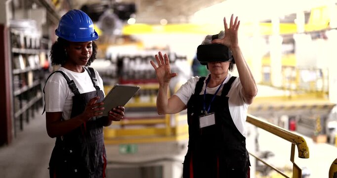 Senior industrial engineer wearing vr headset for training purposes in a modern warehouse, while her younger colleague uses a digital tablet to monitor the simulation progress and provide guidance
