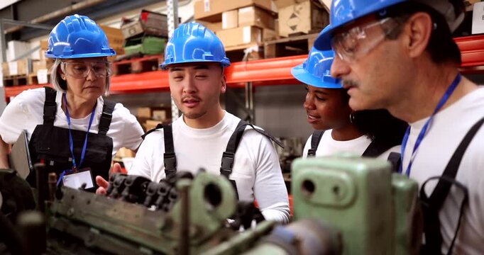 Multiethnic group of engineers wearing hard hats and safety glasses gathering around a machine, discussing its mechanics and operation during a training session inside a large industrial factory