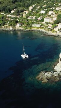 Aerial view of a sailboat anchored in clear blue waters near the rocky coastline and village of Erbalunga, Corsica. Lush greenery and Mediterranean houses line the shore.