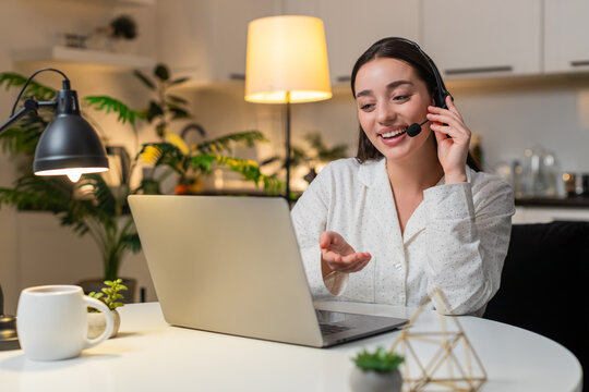 Young woman at night home works as call center agent with headset answers support call and logs ticket. Caucasian girl on sofa at evening listens speak note request polite tone service aid quick log.
