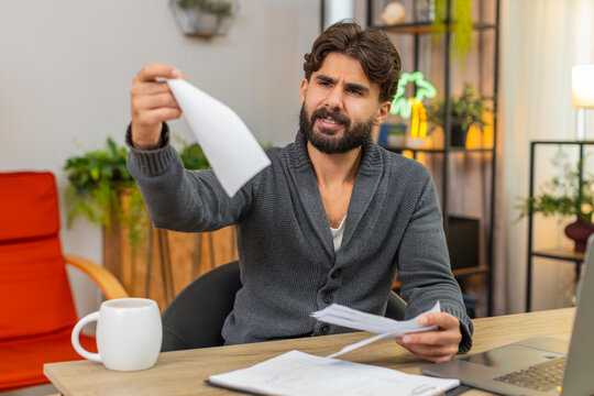 Indian man at home sees unexpected bill feels angry tosses receipt aside no approval gesture at all. Arabian guy at desk checks charges finds extra fee plans dispute calls bank cancels payment quickly