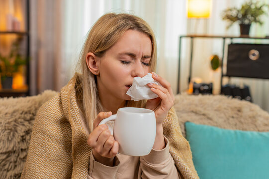 Young ill adult woman sits on home sofa under blanket, holding cup of hot tea and warming hands, sniffling gently. Blonde girl looks tired and sick, reacting to cold symptoms with calm expression.
