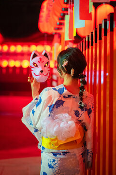 Rear view of a woman in yukata holding a fox mask standing in front of red lanterns at Fushimi Inari Taisha at night. Kyoto, Japan.