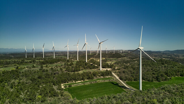 Drone aerial view of multiple wind turbines on green hills forming a wind farm in rural landscape under blue sky.