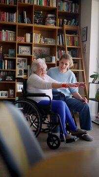 Daughter helping elderly mother in wheelchair with physiotherapy exercise at home.