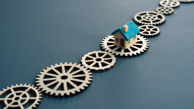 Miniature house model resting on a path of wooden gears depicting the real estate market or home construction process.