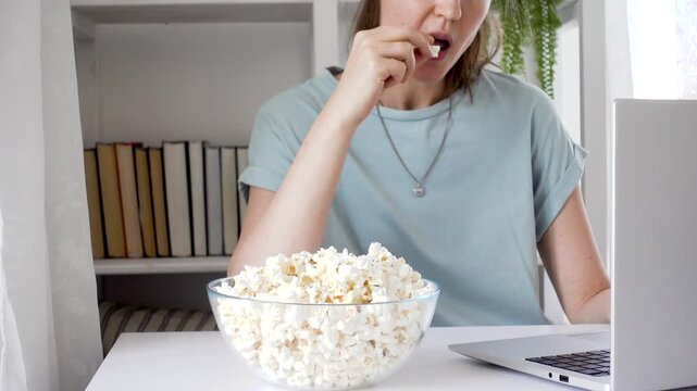 Woman in a blue t-shirt watches a movie on a laptop at home with popcorn. Cozy atmosphere: books, plants, and lighting. Modern home entertainment blends technology and tranquility