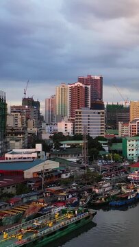 Colorful boats docked along the Pasig River with high-rise buildings and construction cranes in the Manila cityscape under a dramatic sky.