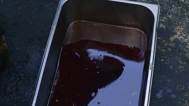 Chef pouring dark pilaf oil from a large ladle into a metal container, close-up