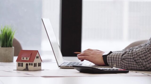 Hands calculate costs near a miniature house. Laptop sits beside a tiny home model. Focused expression suggests financial planning. Ideal for real estate, mortgage, or budgeting concepts