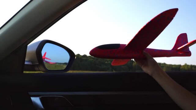 Hand holds red foam airplane near car window. Reflection shows plane in side mirror. Sunlight shines through open car window. Person enjoys flying toy during drive. Perfect for travel, adventure