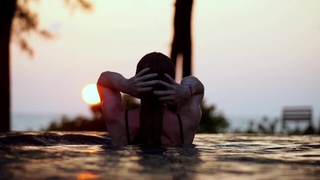 Sunset glows behind a woman in water. She holds her hair as she relaxes. Silhouettes of trees frame the scene. Warm light bathes the pool's surface. Emotion is calm, tranquil, and serene