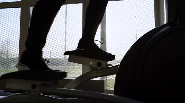 Silhouetted legs move rhythmically on a stepper machine. Bright windows frame the workout routine. Focus remains on motion and fitness discipline. Ideal for health, wellness, or gym activity visuals