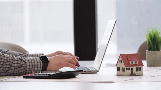 Hands type on laptop, focused, professional. Mini house model sits beside keyboard, symbolizing real estate. Green plant adds natural, calm atmosphere. Use for finance, real estate