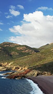 Scenic aerial view of the rugged coastline and hills at Delta du Fango in Corsica, with waves crashing along the rocky shore under a bright blue sky.