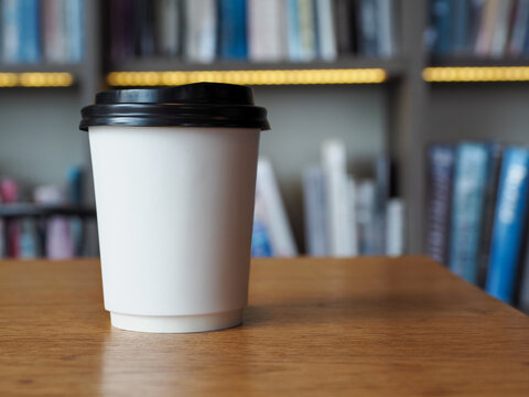 Paper cup coffee cup with black lid on wooden table in blurred library background, minimalist takeaway drink scene