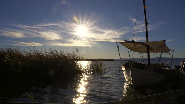 A picturesque wooden pier and a traditional boat moored, used for tourist excursions, in the Albufera Natural Park, with the sun's rays filtering through the clouds and illuminating the calm waters.