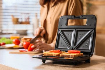 Modern sandwich maker with snacks against woman cutting tomato on table in kitchen, closeup