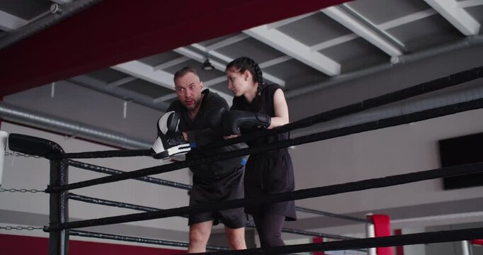 A trainer explains training aspects to a young female boxer while standing in the ring. He analyzes the training process and addresses mistakes for further progress. Slow motion 4k footage.