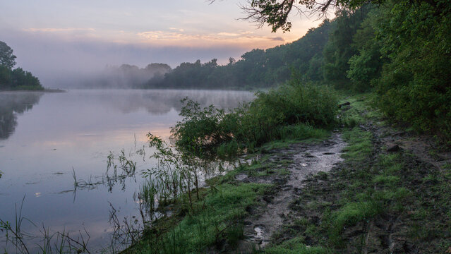 A misty sunrise over the river. Green banks with a muddy path. Peaceful nature scene